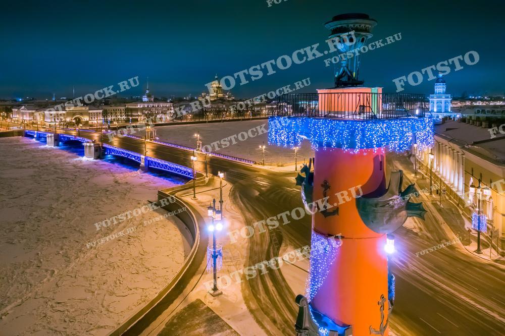 На фотографии изображена Стрелка Васильевского острова в Санкт-Петербурге зимней ночью. Ростральные колонны украшены новогодними гирляндами, создавая праздничную атмосферу. Замерзшая река Нева и подсвеченный Дворцовый мост добавляют особый шарм зимнему пейзажу. Вдалеке виден Исаакиевский собор и другие исторические здания.